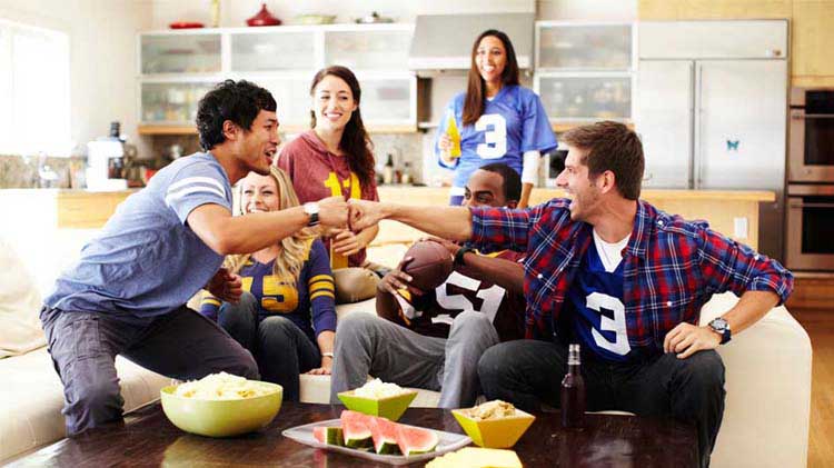 Football fans watching the big game and fist bumping on a couch with snacks.