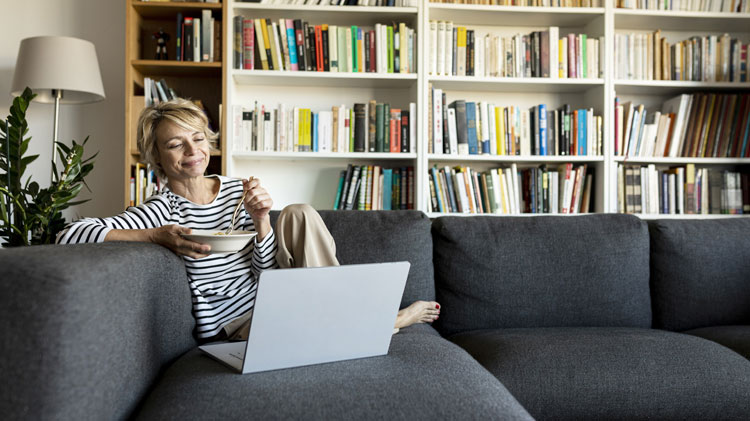 Mature woman using a laptop to work on her digital estate plan and having lunch on a couch.