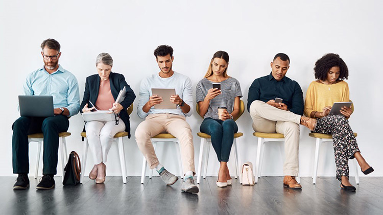 Six people sitting in a row of chairs looking at their laptops, smartphones and tablets.