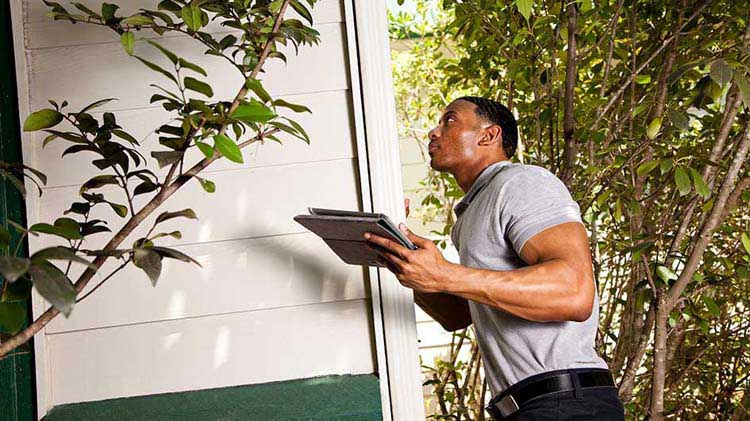 Man looking up holding a tablet while inspecting the exterior of a home.