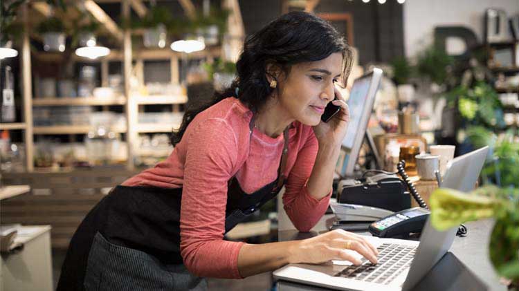 Woman who owns a small business start-up is on her cell phone in her flower shop typing on a laptop.