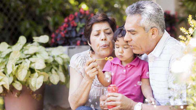 Grandparents with their young granddaughter sitting outside in a garden blowing bubbles.