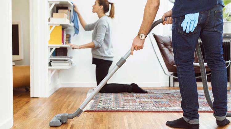 Woman dusting a bookshelf and man vacuuming to get rid of dust in the home.