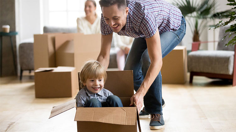 Father pushing son across the floor in cardboard box with mother and other moving boxes behind them.