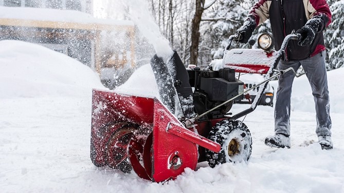 Person walking behind a snowblower as they are clearing the pavement.