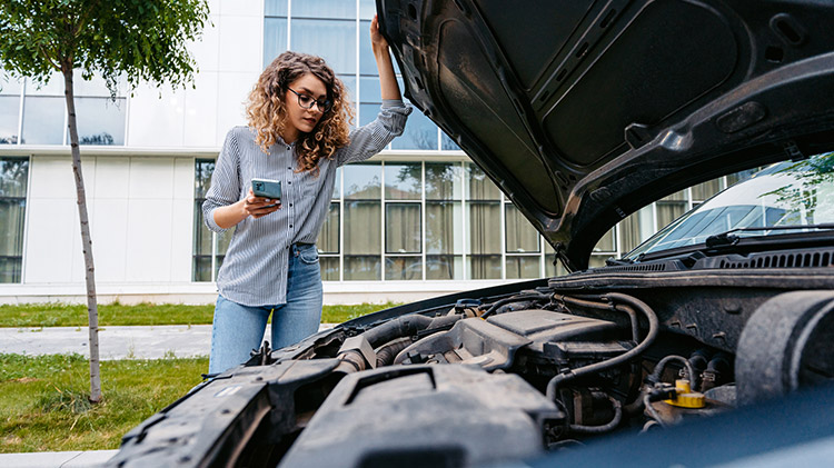 Young woman calling road assistance because of car problems while looking under the hood of the car.