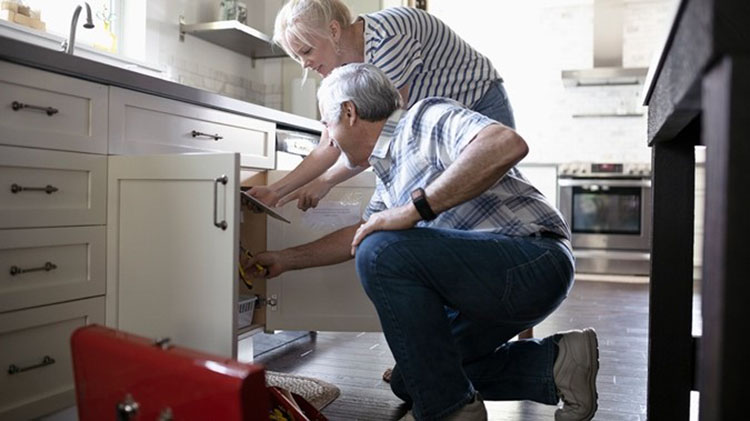 Man kneeling in front of the kitchen sink reading instructions a woman is holding. A toolbox is open beside the man.