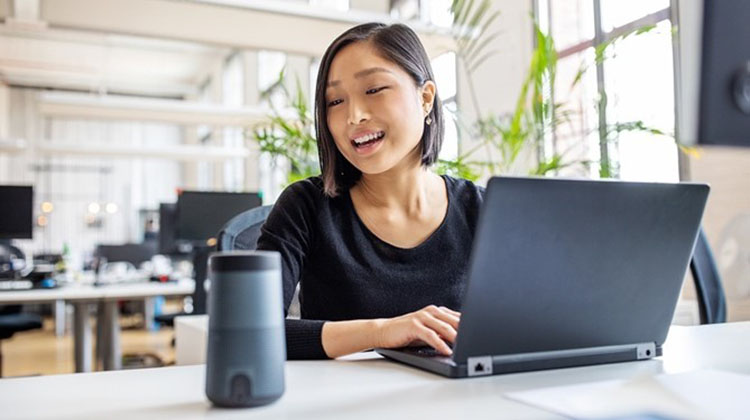 Young woman interacting with a digital voice assistant while working on her laptop.