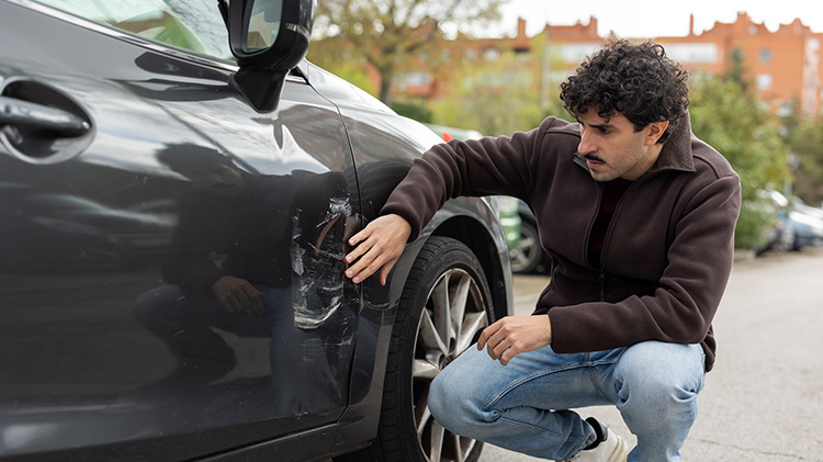 A driver kneels next to his passenger side door, inspecting the damage done by another driver’s vehicle.