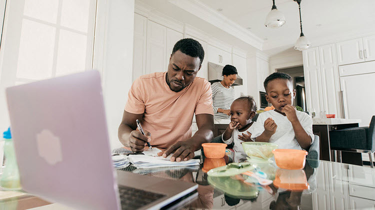 A dad working at a kitchen table while kids eat and the mom is in the background.