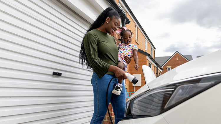 Young woman plugging in her electric vehicle outside her garage while holding a little girl on her hip.