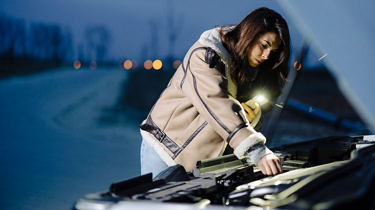 Young woman checking her engine with her cell phone light on a dark road while calling for roadside assistance.