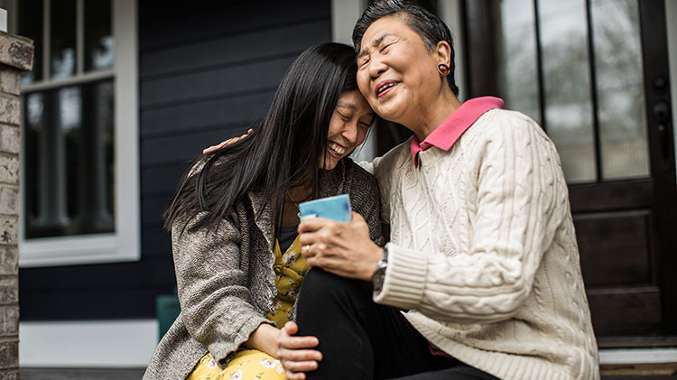 Older and younger woman sit together on a porch, leaning into one another and laughing