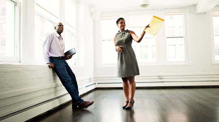 Man and woman standing in an empty apartment with dark hardwood floors and large windows along two walls..