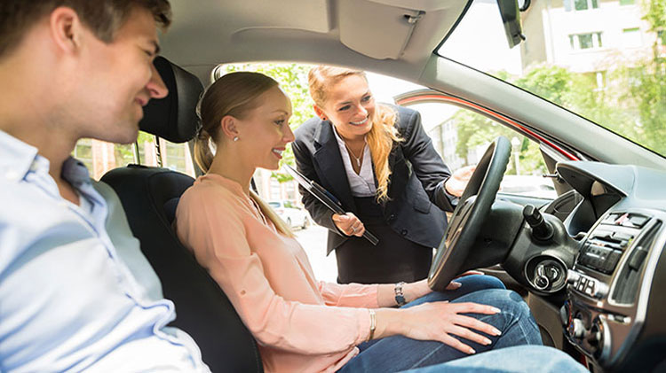 A couple is sitting in a car with the salesperson looking in the window and discussing leasing or buying the vehicle.