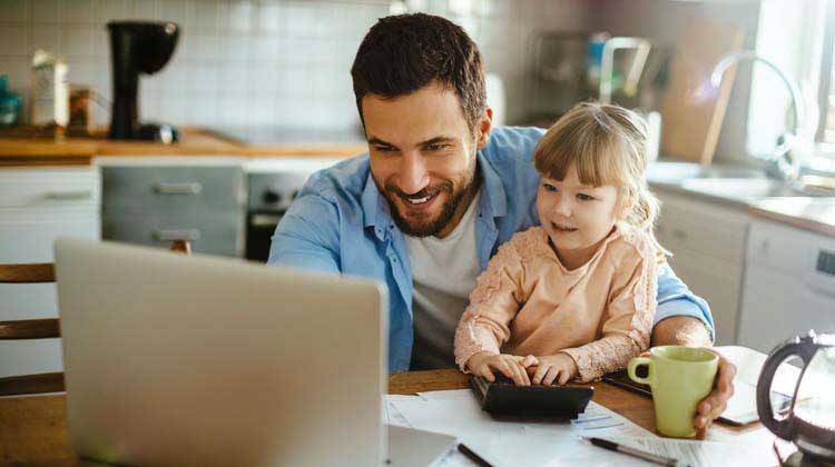 A man with his daughter sitting on his lap at the kitchen table working on a laptop exploring reasons to save money and the different kinds of savings accounts available.
