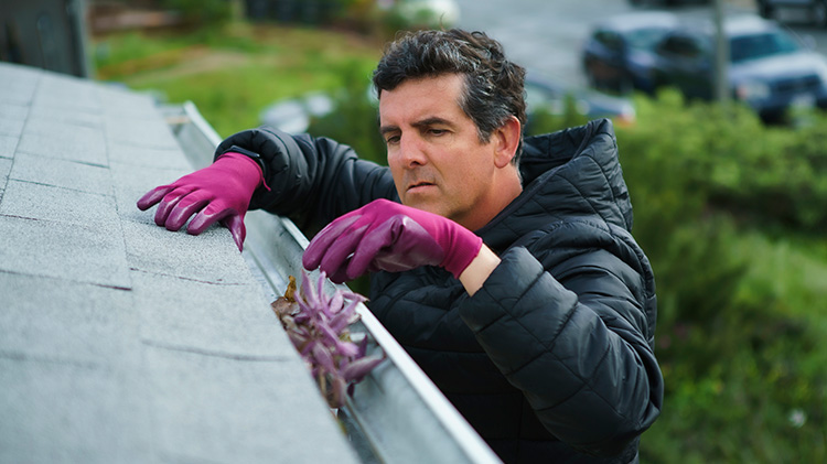 Man performs roof maintenance by cleaning gutters of a roof while wearing gloves.