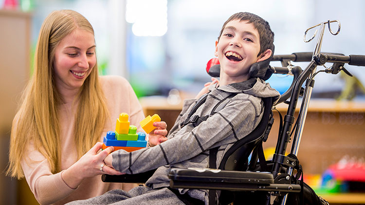 A boy in wheelchair smiles as a woman hands him toy blocks.