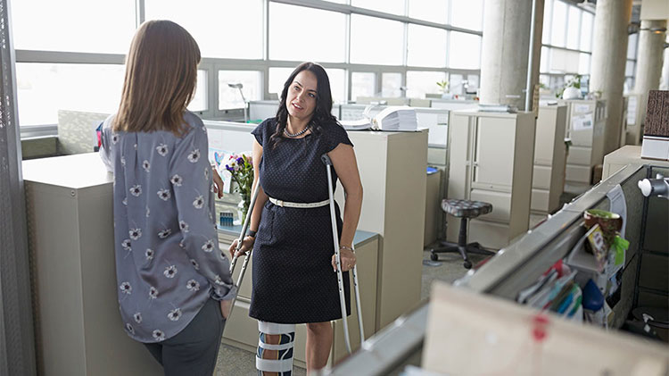 Two women standing in office and one has a brace on her leg and is using crutches.