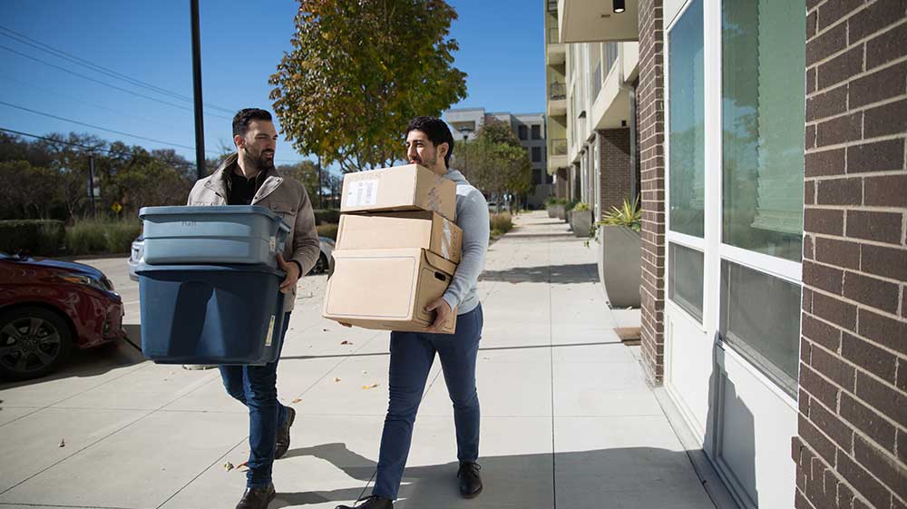 Two poeple holding moving boxes and containers, walking down a sidewalk in front of an apartment building.