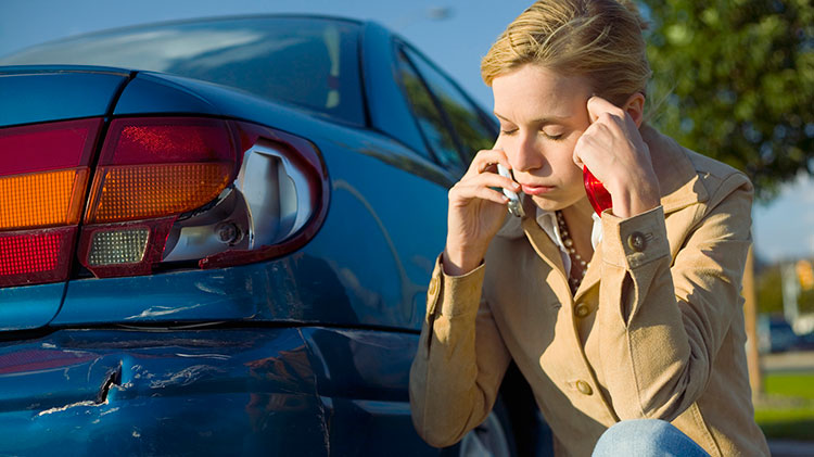 Frustrated woman on phone inspecting damage to car bumper after a not-at-fault accident.