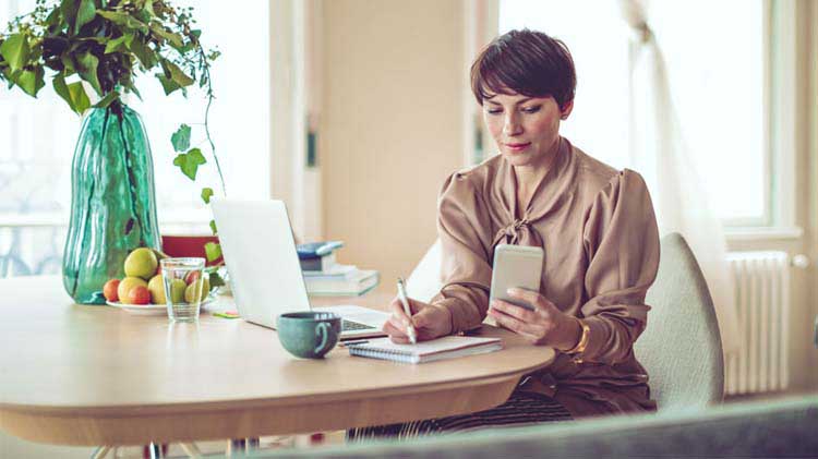 Woman sitting at a table, writing on paper while using her phone and laptop to research money market accounts.