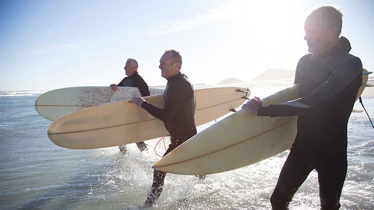Three retired men are entering the ocean to surf.