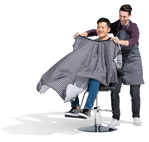 A young, male barber puts a striped cape over a male customer, sitting in the barber chair.