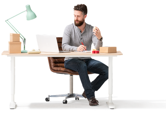 Un joven de raza blanca de barba y pelo oscuros está sentado en una silla de cuero marrón, con ruedas, frente a un escritorio de computadora. Lleva puestos una camisa gris y pantalones oscuros. Sobre el escritorio, hay una lámpara verde, dos cajas de madera y una computadora portátil blanca.