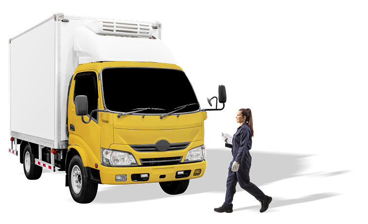 Una joven de raza blanca con el pelo recogido en una coleta alta y vestida con chaqueta, pantalones y zapatos oscuros, y guantes grises lleva en las manos papel blanco. Pasa frente a un camión mediano de caja blanca y cabina amarilla.