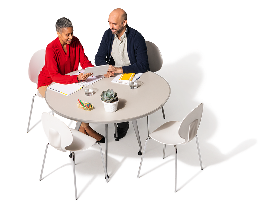 Sentadas una al lado del otro, en una mesa, dos personas revisan una tableta. Hay dos sillas frente a ellos. Sobre la mesa, hay cuadernos, vasos de vidrio, una planta pequeña y marcadores de colores.