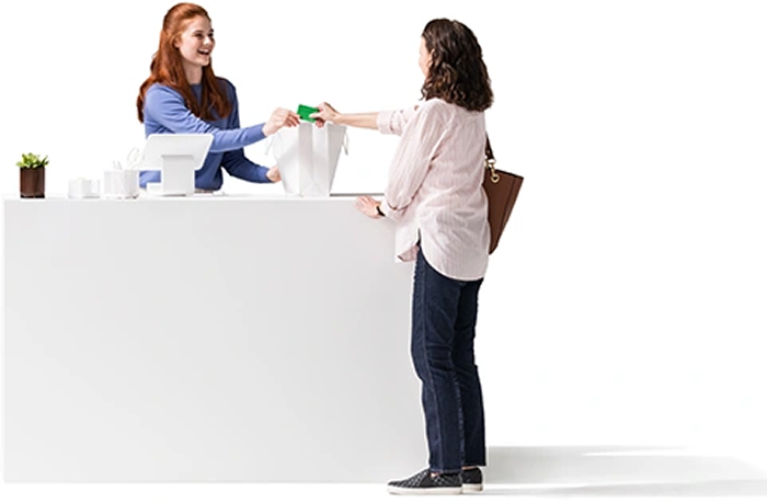 A dark-haired woman, wearing a white shirt, jeans and carrying a purse, hands her credit card to the female shop worker to purchase an item in a white bag on a counter. The young, red-haired shop worker, wearing a blue shirt, stands behind a counter. On the counter is a white tablet on a matching stand, a couple of white containers, and a green plant in a brown pot.