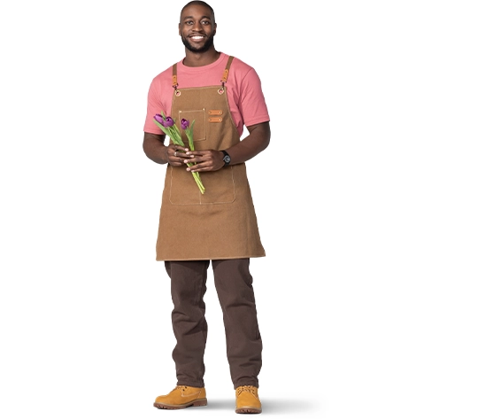 A business owner in a brown apron and khaki pants stands with flowers in hand.