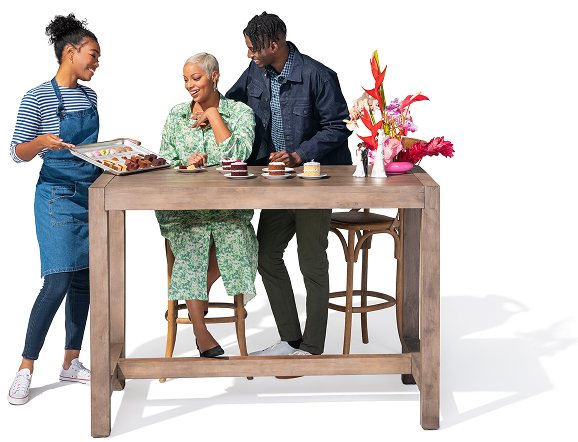 Una joven afroamericana, vestida con una camisa a rayas azules y blancas, jeans, tenis blancos y un delantal azul, muestra una bandeja de pasteles a una pareja afroamericana, sentada en taburetes de mimbre en una mesa alta, que está probando pasteles de boda. A la izquierda de la pareja hay un adorno de novios para el pastel y un ramo de flores rojas.