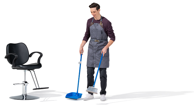 Un barbero joven, caucásico barre con una escoba de mango azul y un recogedor azul. Lleva un suéter negro y jeans, tenis blancos. Está de pie junto a una silla de barbero.