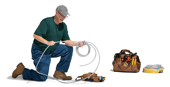 Un trabajador con gorra gris, anteojos, camiseta verde, jeans y botas de trabajo marrones está arrodillado y sosteniendo un cable de fibra. Cerca, en el suelo, hay un cinturón de herramientas, una bolsa de herramientas y más cables.