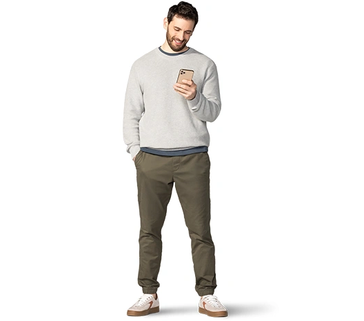 A young, white man, with dark hair, beard and mustache, wears a light, gray sweatshirt, dark gray t-shirt (underneath), olive-green sweatpants, and athletic shoes. He smiles as he looks down at his smartphone.