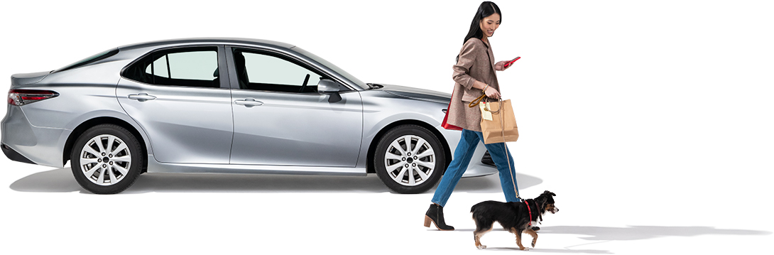 A woman walks her dog past a parked grey sedan.