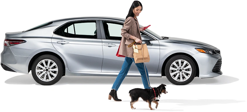 A woman walks her dog past a parked grey sedan.
