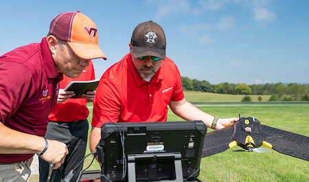Photo is of two State Farm associates, one wearing a Virginia Tech ball cap and the other wearing a dark hat with white logo. They stand before computer device. Man on the right is holding a black drone with yellow markings. The background is lush green fields with stand of trees in the background.