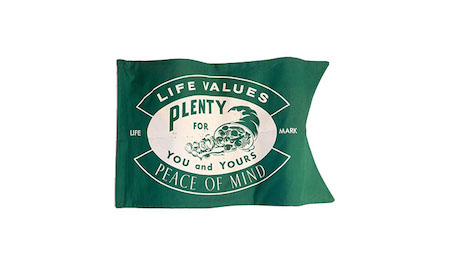 Photo is of State Farm’s 1929 Life Company logo. The white logo appears on a green flag. The logo features a green cornucopia with the words, “Plenty for you and yours,” centered in a white oval. White outlined banners appear above and below the oval. The top one contains the words “Life values” and the bottom one has “Peace of mind.”