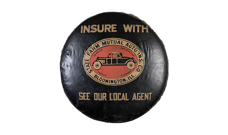 Photo is of a circa 1931 tire cover. The cover is black with a gold-red-and-black State Farm logo, featuring a car. Gold lettering above the logo reads “Insure with,” and text below the logo reads 'See our local agent'