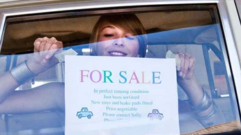 A smiling young lady places a For Sale sign in her car window.