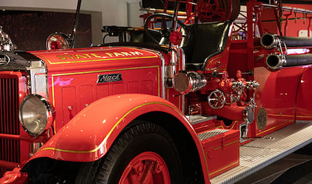 Picture is of a rare 1935 Mack Pumper Fire Engine. The restored fire truck is red with black and silver equipment. It has gold trim and lettering that spells out “State Farm.” There’s a black “Mack” logo plate above the driver’s side wheel well that features a black tire and red hubcap.rive my car