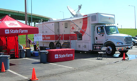 A photo of the Mobile Catastrophe Command Center (M3C) vehicle. This red-and-white truck features a satellite dish. Orange traffic cones mark off the area that features tables, chairs and trash cans. Red-and-white State Farm logo and lettering appear on the vehicle, tent and table drape.
