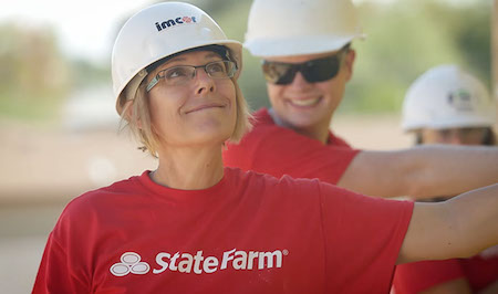 La fotografía muestra a dos empleadas de State Farm, ambas llevan camiseta roja con logotipo de la empresa y casco, ambos de color blanco. La mujer que aparece en primer plano tiene pelo rubio, corto y usa gafas. La otra mujer lleva el pelo recogido y usa lentes de sol oscuros. Ambas mujeres están sonriendo.