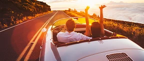 A couple ride into the sunset on a two-laned highway. The man drives the convertible, while the woman raises her hands, joyously, in the air.