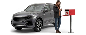 Una mujer joven con una chaqueta prieta y vaqueros de color azul oscuro parada entre su buzón de correo y un SUV gris revisa su teléfono celular inteligente.