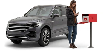 Una mujer joven con una chaqueta prieta y vaqueros de color azul oscuro parada entre su buzón de correo y un SUV gris revisa su teléfono celular inteligente.