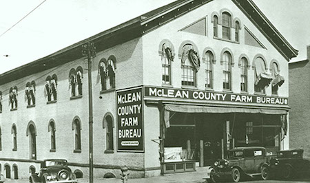 A black-and-white photo of the McLean County Farm Bureau two-story building. Large signs on the front and side with the bureau’s name. Three 1920s-era automobiles can be seen on the street in front of the building.